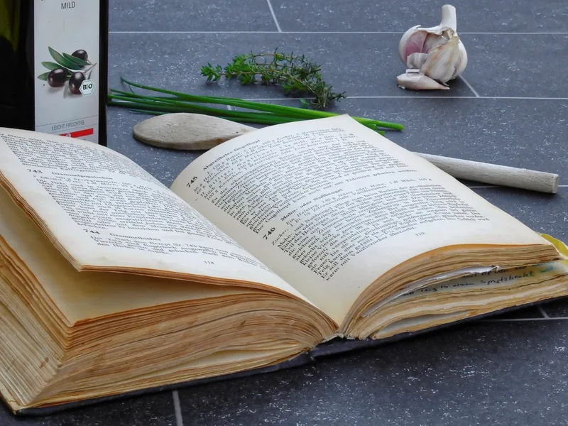 An open cookbook on a kitchen table with herbs and garlic