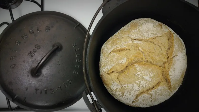 A loaf of crusty no-knead bread inside a black cast iron Dutch oven, with its lid beside it
